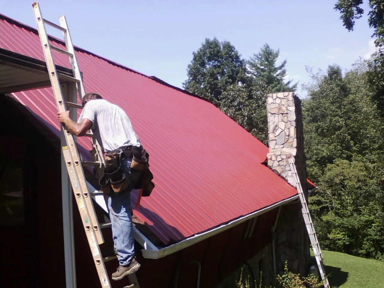 Roofer on a ladder beside a red metal roof and stone chimney — Soffit & Fascia Repair by Roofing Brothers in Fincastle